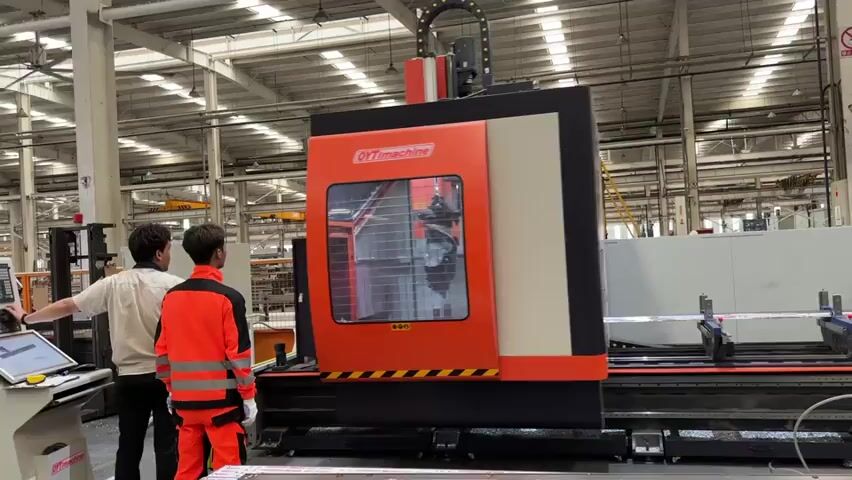 Two men stand in a large factory setting, observing a large orange and black "QYTimachine" milling machine. The man on the left is wearing a white shirt and dark pants, while the man on the right wears an orange safety suit with reflective stripes. The machine is cutting a long, thin metal piece. The factory has a high ceiling with visible support beams and overhead cranes. There are other machines and equipment in the background, creating a busy industrial atmosphere.
