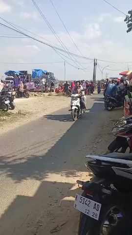 This video shows an outdoor scene on a sunny day with a clear sky. A paved road runs through the middle of the frame, with a dirt shoulder on the left. On the left side of the road, there are several parked motorcycles and a large purple truck decorated with lights and sound equipment. A group of people are gathered around the truck. Further down the road, there are more people and several more parked motorcycles. On the right side of the road, there are more parked motorcycles and a vendor selling colorful snacks. The video primarily focuses on the activity on the road, with several people riding motorcycles. One motorcycle in the foreground is ridden by a man and a woman, both wearing casual clothing. Another motorcycle further down the road is ridden by a woman wearing a hijab and a child. The overall impression is a lively outdoor gathering or event.