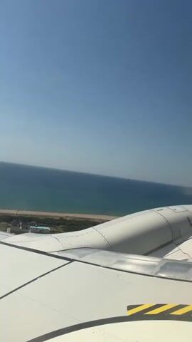 The video captures an aerial view from an airplane window, overlooking a coastline. The primary subjects are the airplane's wing and engine nacelle, dominating the foreground with their white and gray metallic surfaces. A yellow and black striped warning decal is visible on the wing.

Beyond the wing, a vast expanse of the Mediterranean Sea stretches to the horizon, its deep blue waters meeting a clear, bright sky. The shoreline is visible, featuring a strip of sandy beach, some green foliage, and hints of buildings or structures, possibly a resort, nestled near the coast. The scene is bathed in bright sunlight, suggesting a clear and warm day, likely midday given the intensity of the light.

The activity taking place is simply the airplane in flight, cruising over the coastal area of Viladecans, Spain. The perspective from the window implies passengers observing the scenery. The overall mood is serene and picturesque, offering a glimpse of a beautiful coastal landscape from a unique vantage point.

Visible text includes "NO ST" on a metallic plate on the wing, likely part of a warning or identification label. The timestamp at the beginning indicates the video starts at 00:00.