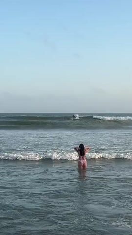 A woman in a pink bikini stands in shallow ocean water facing away from the camera. Waves crash around her. In the distance, a surfer rides a wave. The sky is clear and blue.