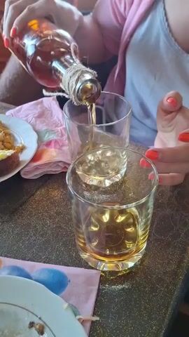 A video shows a person pouring amber liquid from a glass bottle into two glasses. The bottle is tied with twine around its neck. The glasses are on a dark, speckled table. A plate with food, including what appears to be corn, is on the left. In the background, a person in an orange t-shirt and another person in a pink cardigan over a gray tank top are partially visible. The focus is on the action of pouring the liquid.