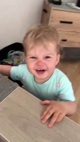 A baby with blonde hair and a light blue shirt with pink flowers is standing and looking at the camera. The baby has a happy expression and is smiling. In the background, there is a wooden dresser and a floor. There are also some toys on the floor. The baby appears to be in a home environment.