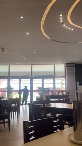 A person stands silhouetted in front of a set of glass doors, which appear to be the entrance to a dining establishment. The doors are partially obscured by blinds. Inside, tables and chairs are arranged for seating. The ceiling features decorative lighting fixtures. Several potted plants are visible near the entrance. To the right, a metal object, possibly a pitcher or container, rests on a table. The overall lighting suggests an indoor environment, possibly during daytime, with natural light filtering through the entrance.