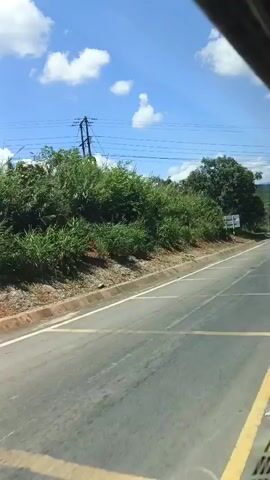The video shows a view from inside a vehicle, likely a bus or van, traveling along a paved road. The road has a white line marking the lane and yellow markings on the asphalt. To the left of the road is a slope covered with dense green vegetation and dirt. A utility pole with wires is visible on top of the slope. The sky is blue with scattered white clouds. To the right of the road, there are more trees and vegetation. A white signpost with red and white striped poles is visible. A dark-colored pickup truck is seen passing by on the road from the left to the right. The camera is slightly tilted, suggesting the vehicle is moving.