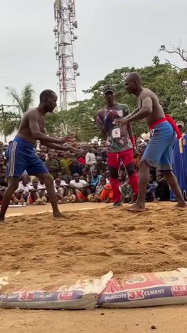 Two shirtless men in shorts are wrestling in a sand pit. A crowd of people surrounds the pit, sitting and standing. A man in a red outfit stands between the wrestlers, acting as a referee. In the foreground, there are two bags of cement marking the edge of the pit. A tall cell tower is visible in the background.