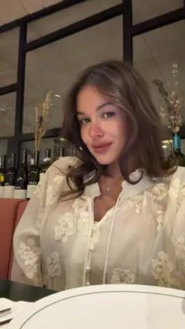 A woman sits at a restaurant table. She has brown, wavy hair that she touches with her hands. She is wearing a white, floral-patterned blouse. Behind her are several wine bottles lined up against a window. A plate and silverware are on the table in front of her. She looks at the camera and smiles.