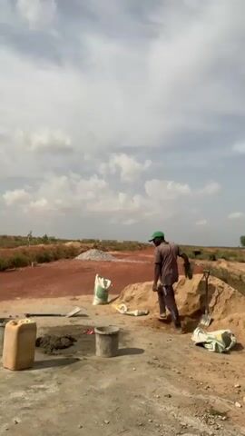 In a medium shot taken in daylight, a man walks and then bends over to set down a bag at a construction site. The man is wearing dark pants, a long-sleeved, dark-colored shirt, and a green baseball cap. He is barefoot. In the foreground is a tan plastic container, a pile of what appears to be mud, a metal bucket, and more bags similar to the one the man is carrying. Behind these objects is a pile of sand with a shovel leaning against it. Further back, there is a pile of rocks and the ground is a reddish color. In the distance, the scene opens up to a wide view of flat land with sparse vegetation. The sky is mostly blue with patches of white clouds.