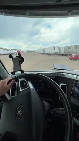 The video shows the interior of a truck cab, looking out the windshield. A person's left hand is visible on the steering wheel. The truck is parked in a lot filled with rows of white semi-trailers. The ground is dirt or gravel. The sky is cloudy. The truck's dashboard is visible, including the steering wheel with a logo in the center. A phone mount with a phone is attached to the windshield. The truck appears to be stationary.
