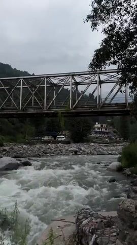 A wide, turbulent river flows from left to right, its whitecaps churning over rocks. A metal truss bridge spans the river in the upper portion of the frame. The banks of the river are rocky, with some vegetation visible. In the background, trees cover a hillside, and a few buildings can be seen near the bridge. The sky is overcast.