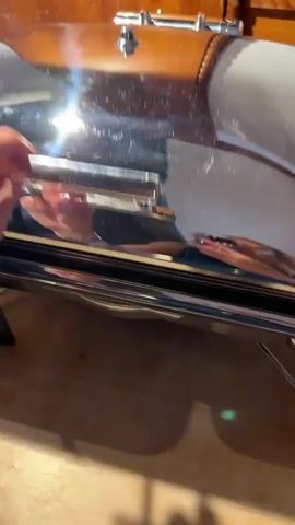 The video pans across a buffet serving station. In the first frame, whole brown eggs are displayed in a silver chafing dish filled with what appears to be sand or coarse salt, with a small black label in the sand reading "hart". The camera then moves to the right, revealing two more chafing dishes. The left dish contains cooked bacon labeled "Speck Bacon". The right dish contains scrambled eggs labeled "RÜHREI Scrambled Eggs". A used spoon with remnants of scrambled eggs sits in a small white dish in the foreground.