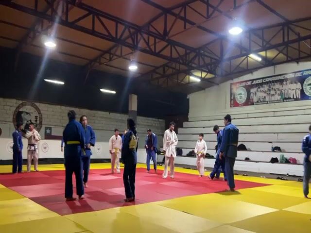 The video shows a group of people practicing judo in a gymnasium. The floor is covered with red and yellow mats. There are several people wearing judo uniforms, some in blue and some in white. Some people are standing and talking, while others are practicing moves. There are also benches on the side of the room, and some bags and equipment are visible. The room is well-lit with overhead lights. There are posters on the wall, including one that says "CLUB DE JUDO CARABINEROS DE CHILE".