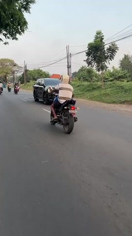 The video shows a rear-view of a person riding a black motorcycle on a paved road. The rider is wearing a light-colored helmet and a striped shirt. In front of the motorcycle, a black SUV is traveling in the same direction. The road appears to be a two-lane road with other motorcycles visible in the distance. The environment is outdoors with trees and vegetation on the side of the road, and utility poles with wires overhead. The lighting suggests it is daytime, possibly late afternoon or early evening. The camera is mounted on the back of the motorcycle, capturing the scene directly ahead.