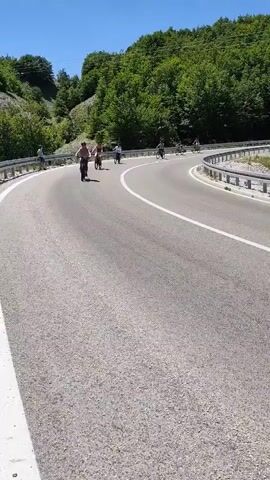 A group of people are riding bicycles on a paved road that curves to the right. The road is bordered by a guardrail on the right and a steep, wooded hillside on the left. The sky is clear and blue. The cyclists are spread out, with some in the foreground and others further back. The person in the immediate foreground is riding a black bicycle and is wearing a helmet and a pink top. The person behind them is wearing a white top. Further back, other cyclists are visible, also on bicycles. The environment appears to be a mountainous or hilly region with lush green trees. The overall scene suggests a recreational activity or a cycling tour in a natural setting.