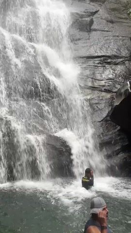A wide shot shows a boy standing in water at the base of a waterfall. The waterfall is tall, spilling down a rocky cliffside. The boy is wearing a swimsuit and appears to be enjoying the experience. To the right of the frame and closer to the camera is a man with a gray hat, who is partially visible. The overall scene suggests an outdoor recreational activity. The water is foamy and the surroundings are rocky, indicating a natural environment.