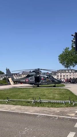 A dark gray military helicopter with the word MARINE and a French flag insignia is on a grassy area. The helicopter has its doors open and a person inside. In the background, there is a clear blue sky, trees, and buildings. A crowd of people are visible in the distance. A traffic light is on the right side of the frame. The foreground shows a road and a decorative fence.