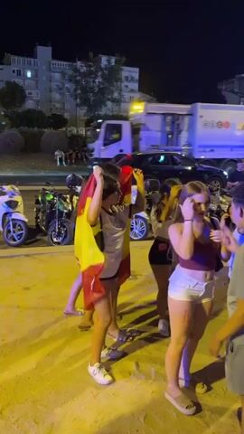 At night, several young women and men are gathered on a sandy area adjacent to a street. One woman is wearing a Spanish flag as a cape. Another woman has face paint on her cheeks. Several motorcycles and scooters are parked nearby. A large white garbage truck is driving down the street in the background. The scene appears to be festive, with small flags strung across the street in the background. The lighting is dim, suggesting it is evening or night.