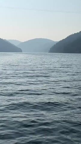 The video shows a wide shot of a large body of water, likely a lake or reservoir, with gentle ripples on its surface. In the background, rolling hills are visible, covered in trees and fading into a hazy blue. The sky is a pale, overcast blue, with a thin wire or cable visible across the top of the frame. The overall impression is one of a tranquil, natural landscape. There are no people or distinct objects visible in the video.