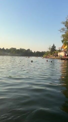 The video captures a wide shot of a lake on a clear day with people swimming in the water and relaxing on the shore. In the foreground, gentle ripples create patterns on the surface of the water. As the view pans to the right, a sandy shore is visible with several people sunbathing and standing. A large tree with lush green foliage stands prominently on the shore, casting a reflection onto the water. Behind the tree, a yellow building with a white roof and balconies can be seen. The background features a dense line of trees, suggesting a natural, wooded environment surrounding the lake. The sky is a clear, pale blue, indicating fair weather. The overall atmosphere appears to be peaceful and recreational, with individuals enjoying a day outdoors.