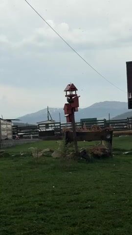 The video shows a rural landscape with a birdhouse on a wooden post in the center. The birdhouse has multiple levels and is painted red.  Behind the birdhouse is a wooden structure resembling a planter or feeding trough, filled with dry material. To the left of the birdhouse is a small, cultivated evergreen tree. The ground is covered in green grass.  In the background, there's a fence and various outbuildings, including what appears to be a greenhouse.  Mountains are visible in the distant background under a cloudy sky, and it is hailing.