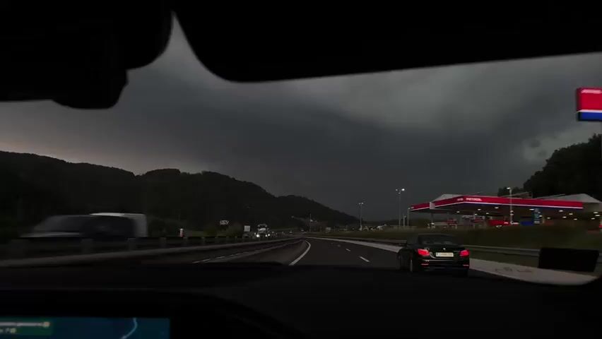 The video captures a view from inside a vehicle driving on a highway. The sky is dark and cloudy, suggesting an approaching storm. In the distance, there are mountains. On the right side of the highway, a gas station with red and white signage is visible. Several vehicles, including trucks and cars, are on the road. At approximately 00:01, a lightning strike illuminates the clouds with a pinkish hue. The camera appears to be mounted on the dashboard or windshield, facing forward.