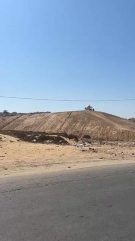 The video shows a large pile of dirt and debris against a clear blue sky. A small structure, possibly a shack or a temporary shelter, is visible on top of the pile. There appear to be people and what might be a vehicle or machinery near the structure. The foreground shows a road with some scattered trash. A utility pole stands to the right of the dirt pile. The scene appears to be an outdoor, possibly rural or industrial, environment.