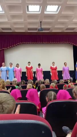 The video shows a group of women walking across a stage in front of an audience of children. The women are all wearing knee-length dresses in pastel colors – blue, pink, red, and lavender, all patterned with white polka dots – plus short white gloves. The children in the audience are sitting in red theater seats and are wearing a variety of colorful clothes, with many wearing pink headscarves. They clap as the women walk past them. The stage has a plain white backdrop, framed with maroon curtains. The ceiling of the auditorium has a grid pattern with embedded lights. A projector is visible above the stage.