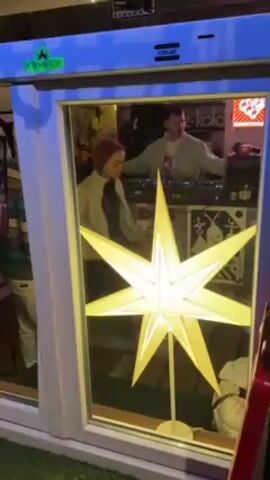 A person in a light jacket and red hair looks into a window while another person wearing a black beanie and gray/blue striped shirt dances outside. Inside the window, a DJ wearing a light jacket and t-shirt mixes music at a table with turntables and audio equipment. A large, illuminated star decoration is positioned in front of the DJ inside the window, adding to the ambient light. A poster of a spiral design is visible behind the DJ.  Other decorations and signage are present, with "fritz-kola" visible. The scene is brightly lit. The window frame is white with blue lighting above.