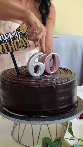 A woman is celebrating her 60th birthday in Santiago Yancuitlalpan, Mexico. She is wearing a light-colored, strapless dress with a floral pattern and has long, dark hair. She is enthusiastically cutting into a rich, dark chocolate cake that is adorned with "Happy Birthday" cake topper and silver and pink candles shaped like the number "60." The cake is displayed on a silver cake stand. The scene appears to be indoors, possibly at a home or a venue, with a table set for a celebration. There are plates, cutlery, and floral decorations on the table. In the background, there's a table covered with a blue tablecloth, stacks of white plates, and bottles, suggesting a party setting. The woman's expression conveys joy and excitement as she cuts the cake. The lighting suggests it might be daytime, and the overall atmosphere is festive.