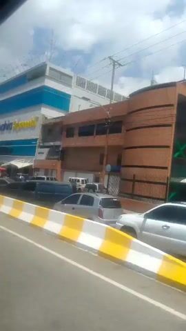 The video captures a view from a vehicle moving past a commercial building. The building has a blue and white facade with large yellow lettering that appears to spell "Bahia Supermarket". A green banner attached to a pole in front of the building reads "LA TIENDA N° 1 DEL PAÍS". Several cars, vans, and motorcycles are parked or driving in the foreground and background. A red tent is visible near the entrance of the supermarket. The sky is partly cloudy.