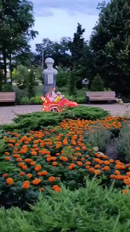 A woman is pushing a child in a small red toy car that resembles a sports car through a park. The child is seated in the toy car. In the foreground, there is a lush garden bed filled with bright orange marigolds and green bushes. Behind the garden bed, a paved walkway is visible, with several park benches, trees, and a statue on a pedestal. A few other people can be seen in the background, some sitting on benches and others walking. A food stand or kiosk is also present. The sky is overcast, suggesting it may be dusk or a cloudy day. In the far background, a Ferris wheel is faintly visible. The overall scene depicts a peaceful park setting with recreational activities.