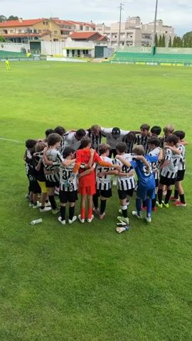 The video shows a group of young soccer players, all wearing black and white striped uniforms, gathered in a circle on a grassy soccer field. They are all looking down, with their arms around each other, in what appears to be a huddle. One player in the center, wearing an orange goalie uniform, is also part of the huddle. There are a few adults standing on the edge of the circle, also looking down. In the background, there are bleachers and buildings. The sky is overcast. The scene is static, with no discernible action or movement beyond the slight jostling of the players within the huddle. The video appears to be a still shot or a very short, unedited clip.