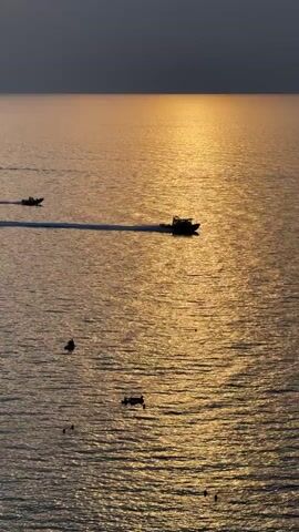 This is an aerial shot of the sea at sunset. A dark sky transitions to a golden horizon, with the sun reflecting on the rippling water. Several boats are visible on the sea. Two boats are moving, leaving wakes. One boat is in the foreground and another is in the mid-ground. Several smaller boats are anchored in the foreground. There are also several people visible in the water in the foreground.
