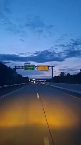 The video shows a view from the driver's perspective of a highway at dusk. The sky is a deep blue with some clouds. There are two overhead signs with directional information. The sign on the left is green and indicates "Seulimeum Seulawah 500 m". The sign on the right is yellow and reads "Akhir Jalan Tol 500 m" which translates to "End of Toll Road 500 m". The headlights of the vehicle illuminate the road ahead, casting a strong orange glow. Other vehicles are visible further down the highway. Trees and vegetation line the sides of the road.