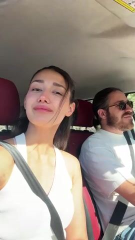 A woman with long brown hair pulled back into a ponytail, wearing a white tank top and a seatbelt, sits in the passenger seat of a car. She looks at the camera and smiles. A man with long brown hair in a ponytail, a beard, and sunglasses, is driving the car. The interior of the car has red seats. The car appears to be moving.