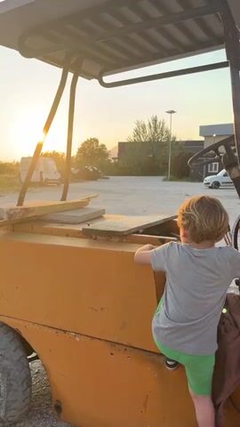 A young boy with short blond hair is climbing onto an old, orange forklift against the backdrop of a setting sun. He is wearing a gray t-shirt, green shorts, and black and white sneakers with socks. He uses the machine's frame and wheel well as steps, holding onto a support bar and the steering wheel as he pulls himself up. The forklift has a sunshade on top and sits on gravel. In the background are trees, a white van, and some buildings under a partly cloudy sky. The sun is low on the horizon, casting a golden light on the scene.
