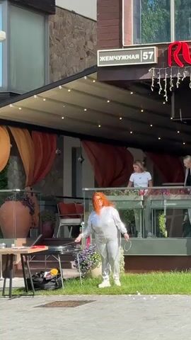 A woman with red hair, wearing a shiny silver jumpsuit and sunglasses, stands on a patch of grass in front of a building with an awning. She is holding a wand with a circular bubble-making apparatus and is surrounded by numerous floating bubbles. In the background, two women are visible on an elevated patio area behind a glass railing. To the left of the woman, a small table with a black bag and some items on it is visible. The building has a sign that reads "ЖЕМЧУЖНАЯ 57" and a red neon sign. The scene appears to be outdoors during the daytime.