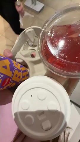 A close-up overhead shot shows a hand holding a McDonald's drink carrier. The carrier holds two drinks, one in a clear plastic cup with a clear lid and a red liquid with ice and fruit, and another in a purple and yellow disposable cup. A white lid sits on top of the plastic cup, and the disposable cup is partially visible. The background is blurred, but appears to be a public space, possibly a fast-food restaurant. The lighting is bright and artificial.