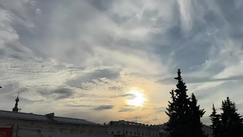 The video shows a view of the sky with clouds and the sun. There are silhouettes of trees and buildings in the foreground. The sun is partially obscured by clouds, creating a halo effect. The sky is a mix of blue and gray, with wispy white clouds. The buildings appear to be old and made of brick, with a dome-shaped structure visible on one of them. The trees are evergreen, and their branches are dark against the bright sky. The overall scene suggests an outdoor setting during the daytime, possibly at sunrise or sunset given the lighting.