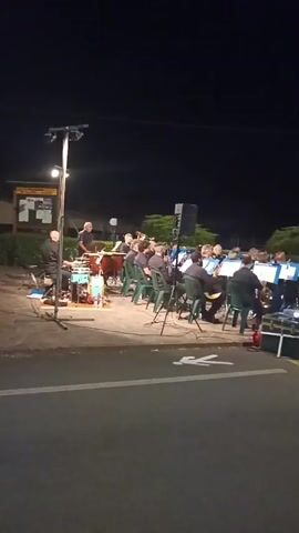 A nighttime outdoor concert is in progress. A band, likely a brass band given the instruments visible, is seated on chairs in rows, performing for an unseen audience. They are illuminated by spotlights and stage lighting. The band members are wearing dark uniforms. In the foreground, a street is visible with a white pedestrian crossing marking. Various pieces of equipment, including speakers, audio equipment, and musical instruments like drums, are set up around the performance area. The background shows a dark sky and some greenery, suggesting a park or public space. The overall atmosphere appears to be a community event or performance.