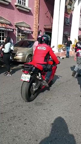 The video shows a group of people and motorcycles on a street. The motorcyclists are wearing helmets and riding red motorcycles with license plates that read “AE2K79N”. Some people are standing and walking, and others are on motorcycles. There is a pink building with white columns in the background, as well as cars and other buildings. Someone is taking pictures with a phone. Some people are wearing masks.
