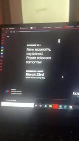 A close-up shot of a laptop screen displays a webpage titled "ECONOMY v1.1". The text on the screen reads, "New economy, explained. Paper releases tomorrow. COMING IN 5 DAYS: MARCH 23rd. More details every day." A graph on the right side of the screen shows a green line trending upwards, with numerical data displayed above. The total USD value is shown at the bottom as $3,727.8. The laptop itself is a Legion model and is positioned on a surface. The background includes a white object with "Projectors" visible on it and a partially visible wall with an electrical outlet. The focus is on the laptop screen and the information displayed.