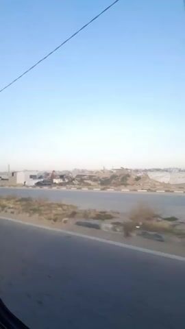 A vehicle is moving down a road on a clear, sunny day. The road has a center divider. The landscape on the opposite side of the road is barren and sandy with sparse vegetation. Further in the distance, there appears to be a shanty town or informal settlement with makeshift structures. A utility pole is visible near the side of the road.