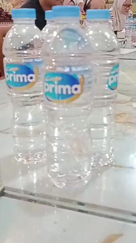 The video shows a close-up, slightly shaky view of several plastic bottles of "Prima" brand water on a table. In the background, blurry figures of people are visible, some wearing headscarves. The setting appears to be indoors, possibly a meeting or gathering, with a patterned wall visible in the far background. The focus is primarily on the water bottles.