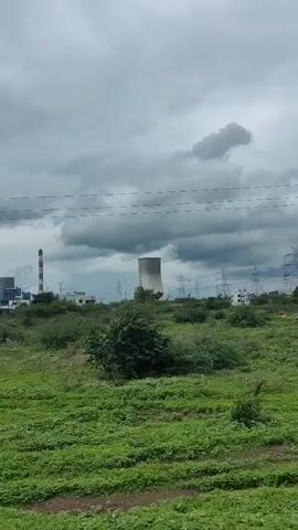 The video shows a view from a moving vehicle, likely a train, looking out at a landscape with a power plant in the distance. The sky is overcast and cloudy. In the foreground, there is lush green vegetation and a single, large tree. The power plant features several tall smokestacks, some emitting steam or smoke, and large cooling towers. Utility poles and wires are also visible. The camera is panning slightly to the right, indicating the vehicle's movement. The overall impression is an industrial facility set against a natural, albeit somewhat bleak, environmental backdrop.