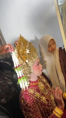 The video captures a close-up, medium shot of a young woman dressed in a traditional wedding outfit, seated and undergoing final preparations. She is adorned in a vibrant maroon dress embellished with intricate gold detailing and a large, ornate gold headdress decorated with white flowers and green accents. Her makeup is prominent, with bold pink blush and lipstick. To her left, a person, mostly out of frame, is seen adjusting her headdress. To her right, another person wearing a light-colored hijab and a brown top looks on attentively, holding a small, light-colored object in their hands, possibly a piece of jewelry or accessory. The background appears to be an indoor setting with a plain white wall on the right and a curtained window or wall on the left. The lighting is bright, illuminating the subjects and their attire. The focus is on the bride and the careful adjustments being made to her appearance.
