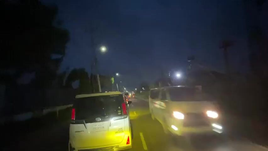 The video captures a night-time street scene from a moving vehicle's perspective. It shows a two-lane road with cars, including a white van and a white sedan, driving in the same direction. There are streetlights illuminating the road and surroundings. The sky is dark, suggesting it's night. There's some vegetation on the sides of the road.