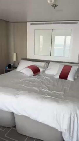 A woman is seen doing a front flip onto a bed. The room appears to be a hotel or cruise ship room with a large bed centered in the shot. The woman is wearing a bright orange athletic top and a black thong. She has brown hair pulled back into a braid. She performs the flip and lands with a slight giggle on the bed.