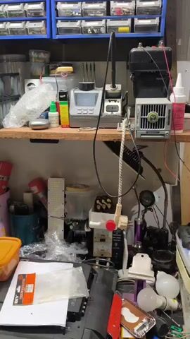 The video shows a workbench cluttered with electronic components and tools. In the background, blue shelves are filled with small transparent drawers containing various items. A wooden shelf above the workbench holds several soldering stations, a roll of bubble wrap, small bottles, and other miscellaneous objects. Below this shelf, the workbench is piled high with more items, including a long LED light strip, various containers, a soldering iron stand, a fan, and electronic equipment. On the desk in the foreground, there are more electronic parts, tools, and a laptop. The overall impression is of a busy workspace dedicated to electronics repair or hobbyist projects.
