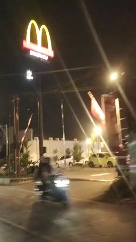 The video shows a nighttime view of a McDonald's restaurant. The iconic golden arches sign is visible and illuminated. Several cars are parked in the lot, and one car appears to be in motion on the road in the foreground. A motorcycle is also visible in motion, adding to the sense of movement. The overall environment suggests a public area with lighting from the restaurant and streetlights illuminating the scene. There are also flags visible, one of which is red and white. The video appears to be recorded from a moving vehicle, indicated by the motion blur and slight changes in perspective.