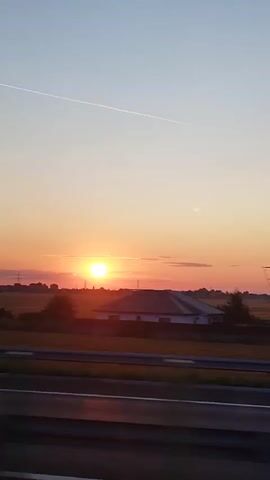 The video captures a rural landscape at sunset, viewed from a moving vehicle. The sky transitions from a soft orange near the horizon to a pale blue above. A sun, low in the sky, casts a warm glow. In the foreground, a highway barrier is visible, indicating the viewpoint is from a car or bus traveling on a road. Beyond the barrier, fields and scattered houses with reddish-brown roofs are discernible. Trees line the edge of the fields. A single contrail streaks across the sky. The overall impression is one of a tranquil journey through the countryside as day turns to night.